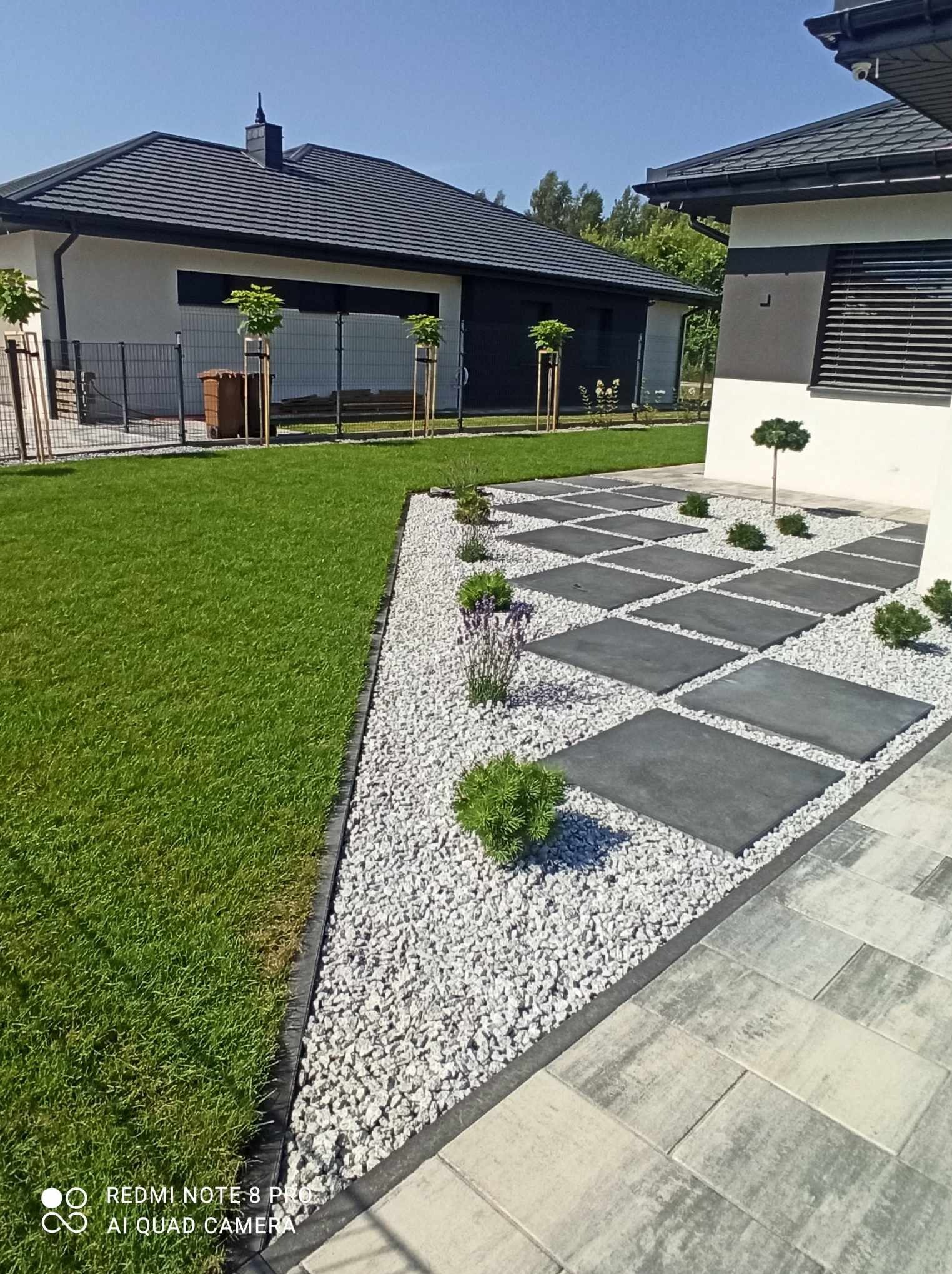 Nowoczesny ogród z równo przystrzyżonym trawnikiem, ścieżką z szarych płyt oddzielonych białym żwirem i młodymi drzewkami przy ogrodzeniu.