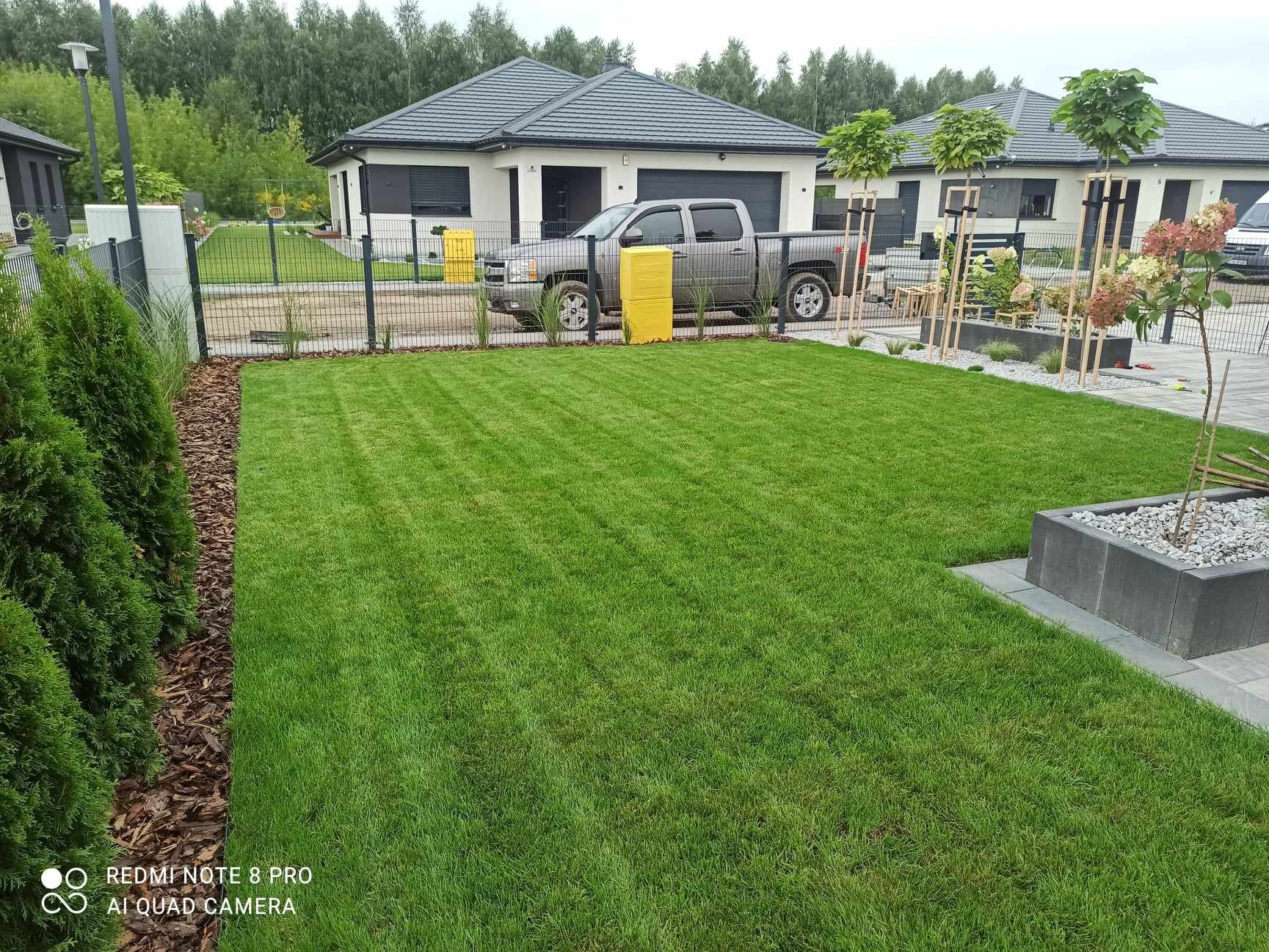 Perfekcyjnie przystrzyżony trawnik przed nowoczesnym domem z szarą ciężarówką na podjeździe, widoczne obrzeża z kory i świeżo posadzone drzewka.