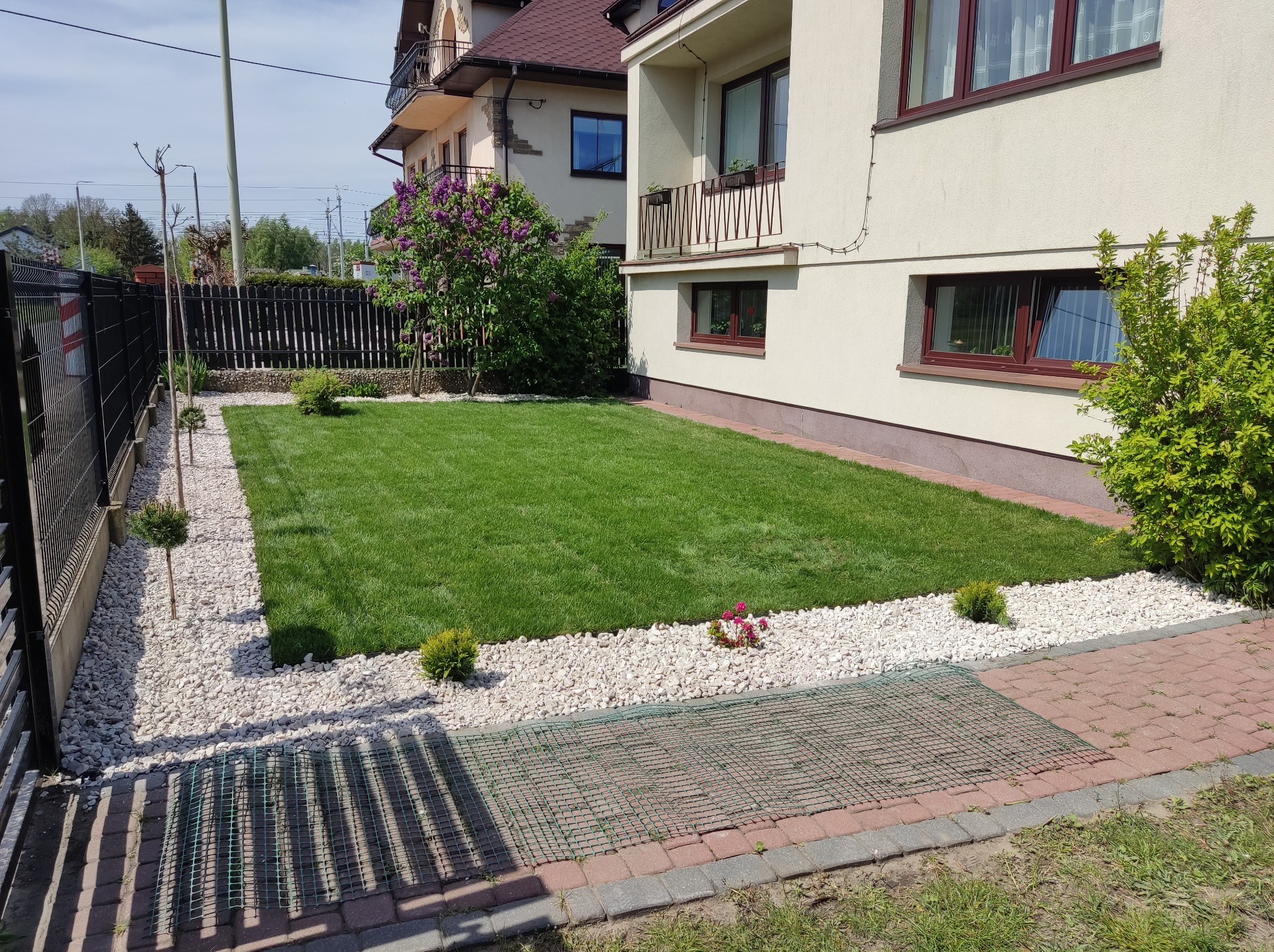 Widok na równo przystrzyżony trawnik, obrzeża z białych kamieni, ozdobne krzewy i drzewka w ogrodzie przed domem z jasną elewacją.