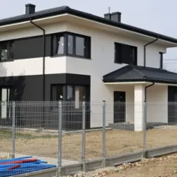 Nowoczesny dom jednorodzinny z biało-czarną elewacją i grafitową dachówką, ogrodzony metalową siatką na betonowej podmurówce, widoczne rury instalacyjne przy ogrodzeniu.