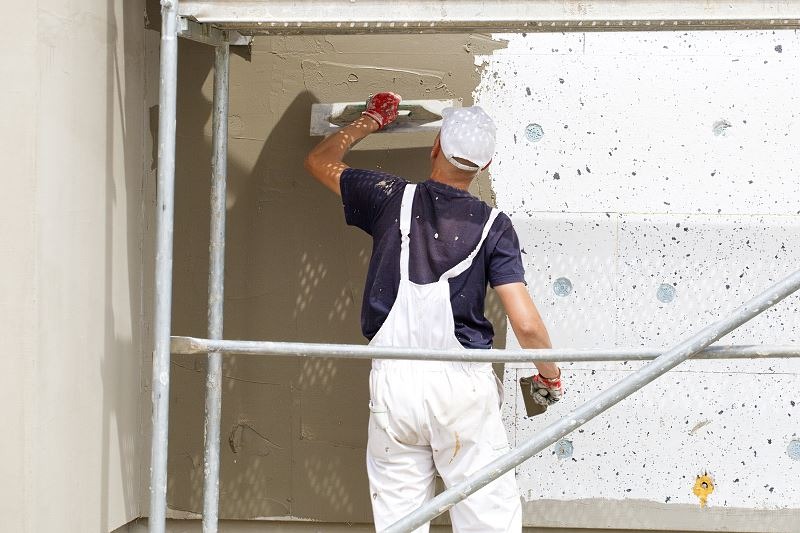 Pracownik w białym kombinezonie, z tyłu, nakłada szarą zaprawę na ocieploną ścianę elewacyjną za pomocą pacy, stojąc na rusztowaniu.