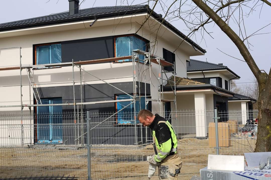 Pracownik w odblaskowej kamizelce przygotowuje materiały na tle domu w Zendek w trakcie prac elewacyjnych, widoczne rusztowania i okna zabezpieczone folią.