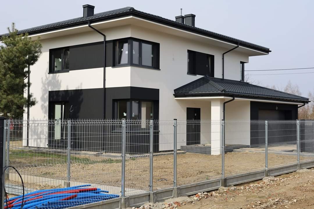 Nowoczesny dom jednorodzinny z biało-czarną elewacją i grafitową dachówką, ogrodzony metalową siatką na betonowej podmurówce, widoczne rury instalacyjne przy ogrodzeniu.