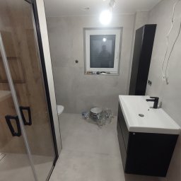 Osoba prywatna - Łazienka w trakcie wykańczania: kabina prysznicowa z drewnopodobnymi panelami, czarna szafka z białą umywalką i czarną armaturą, surowe ściany w odcieniach szarości, widoczne pozostałości po remoncie.