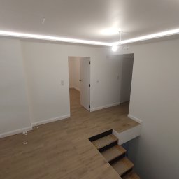 Osoba prywatna - Wnętrze domu po remoncie, widok na drewnopodobną podłogę z płytek, schody z wykończeniem z płytek, białe ściany i sufit z oświetleniem LED, otwarte drzwi do kolejnych pomieszczeń.