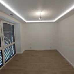 Pusty pokój po remoncie z drewnianą podłogą, białymi ścianami, oświetleniem LED na suficie i dużym oknem z żaluzjami.