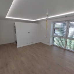 Osoba prywatna - Puste, jasne pomieszczenie z panelami podłogowymi, białymi ścianami, oświetleniem LED w suficie podwieszanym i oknami z żaluzjami, widoczne ślady po pracach wykończeniowych.