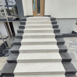 Osoba prywatna - Zewnętrzne schody wejściowe wyłożone jasnoszarymi płytkami z ciemnoszarymi obrzeżami, prowadzące do drzwi wejściowych w nowym budynku, z widocznymi elementami rusztowania w tle.