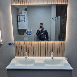 Osoba prywatna - Nowoczesna łazienka z podwójną umywalką, lustrem z podświetleniem LED i drewnianymi panelami. W lustrze odbija się osoba robiąca zdjęcie oraz fragment kotła gazowego.