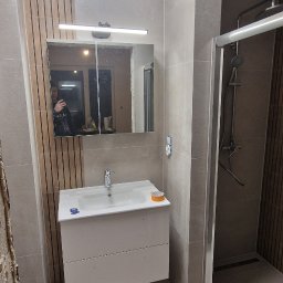 Osoba prywatna - Nowoczesna łazienka w trakcie wykańczania: widoczna umywalka z szafką, lustro z oświetleniem, kabina prysznicowa z deszczownicą, płytki w odcieniach szarości, fragment ściany z drewnianymi panelami...