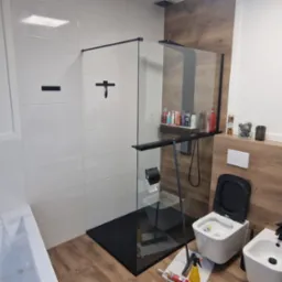 Nowoczesna łazienka z kabiną prysznicową typu walk-in z czarnymi detalami, białą armaturą, obudową z paneli imitujących drewno i częściowo wykończona białymi płytkami. Widoczne narzędzia...
