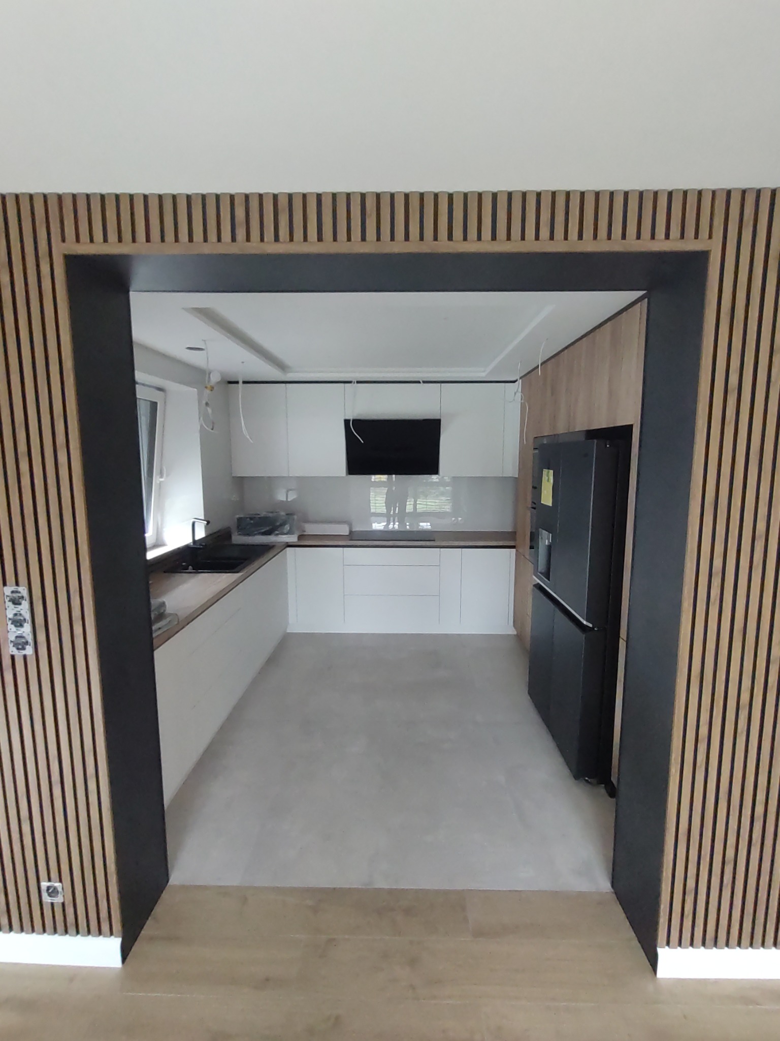 Nowoczesna kuchnia z białymi szafkami, czarnym sprzętem AGD i dekoracyjnymi panelami ściennymi z drewna, widziana z perspektywy wejścia.