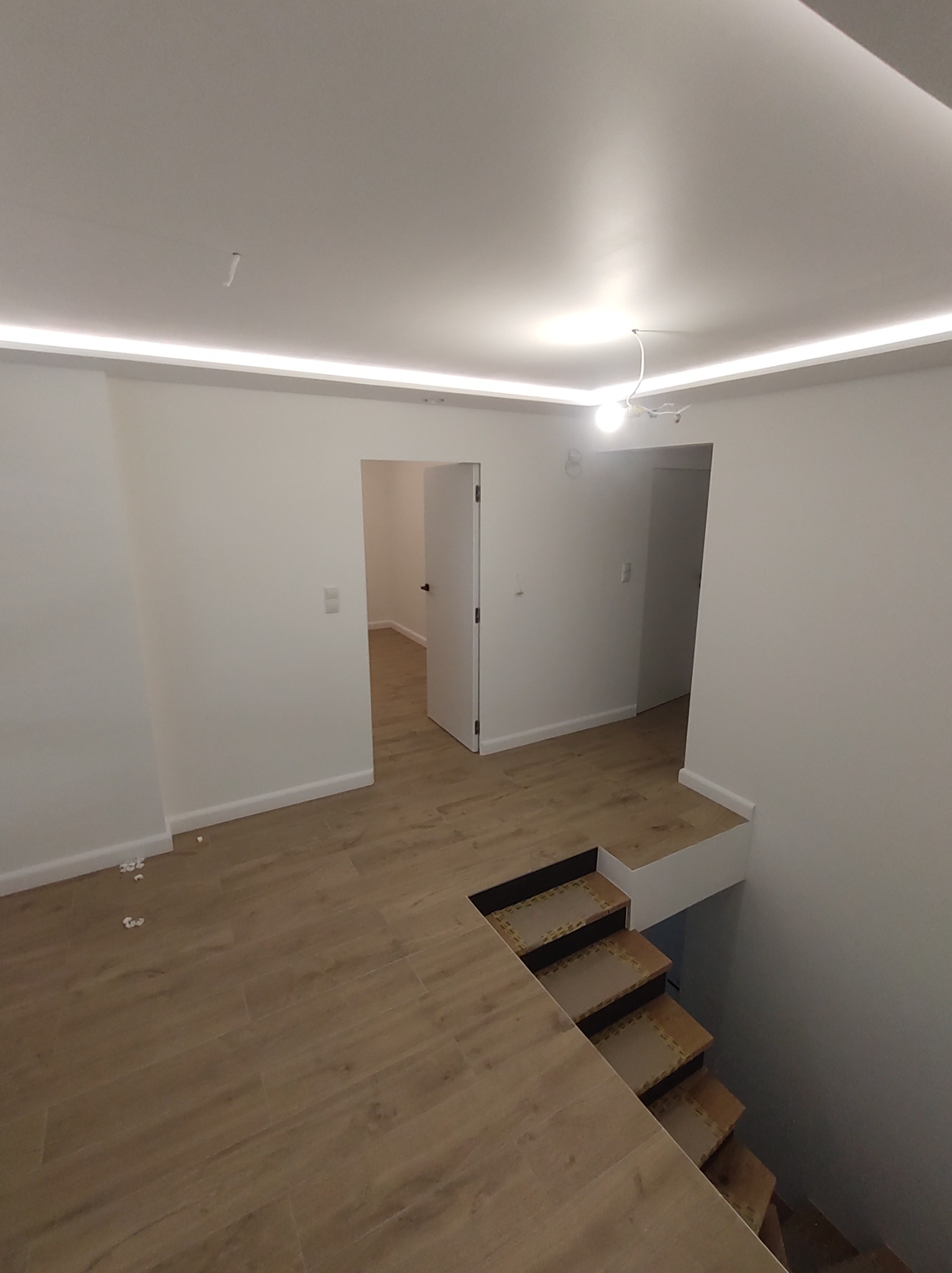 Wnętrze domu po remoncie, widok na drewnopodobną podłogę z płytek, schody z wykończeniem z płytek, białe ściany i sufit z oświetleniem LED, otwarte drzwi do kolejnych pomieszczeń.