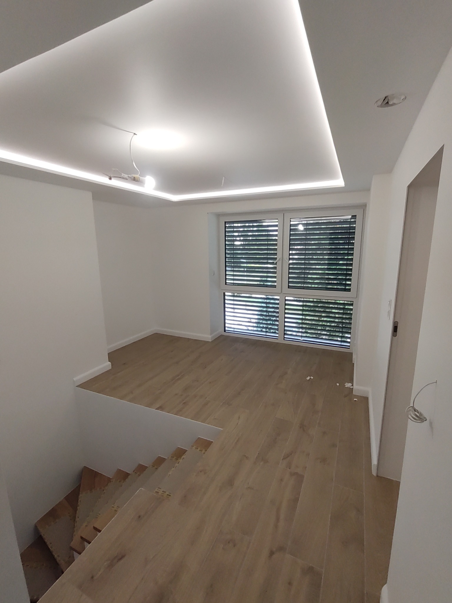Jasne wnętrze z drewnopodobną podłogą, schodami w trakcie wykończenia, oknem z żaluzjami i nowoczesnym oświetleniem sufitowym z listwami LED. Widoczne niedociągnięcia wykończeniowe przy podłodze...
