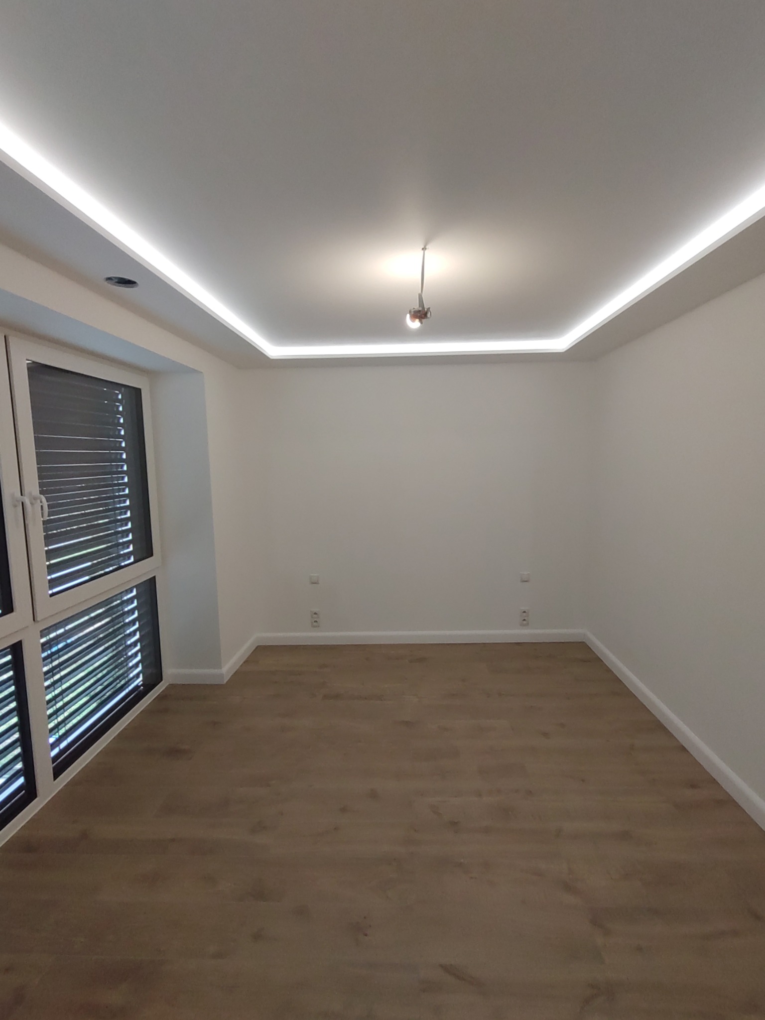 Pusty pokój po remoncie z drewnianą podłogą, białymi ścianami, oświetleniem LED na suficie i dużym oknem z żaluzjami.