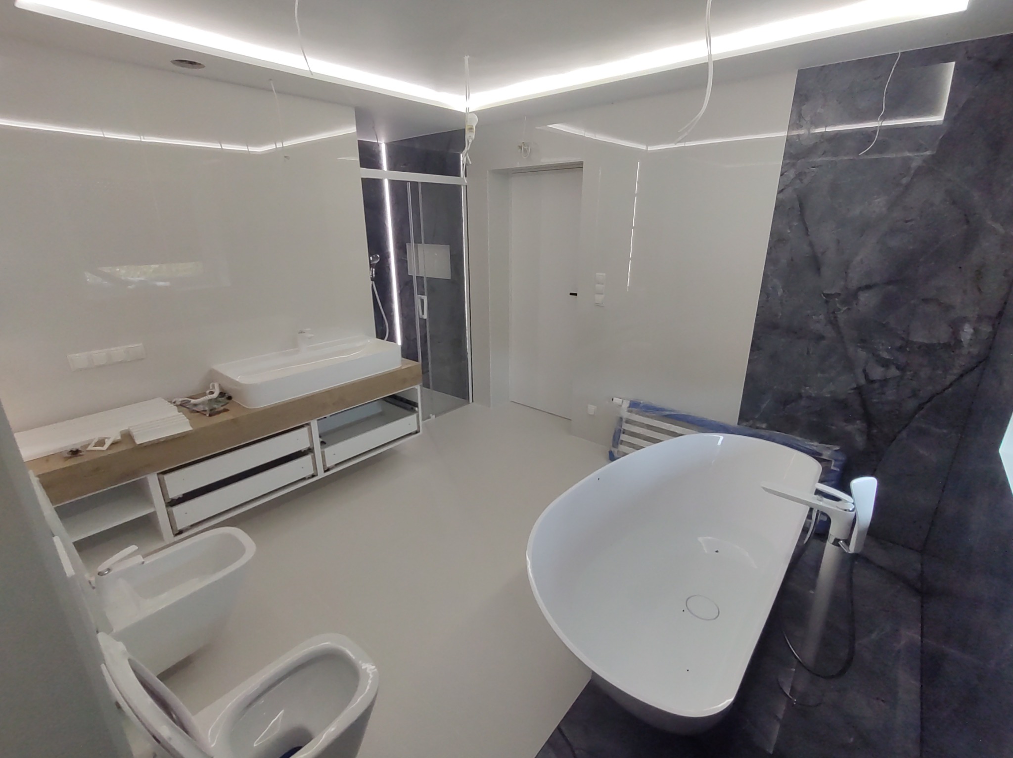 Nowoczesna łazienka w trakcie wykończenia: białe płytki, wolnostojąca wanna, podwieszana toaleta, umywalka nablatowa z drewnianą szafką, oświetlenie LED, widoczne przewody elektryczne.