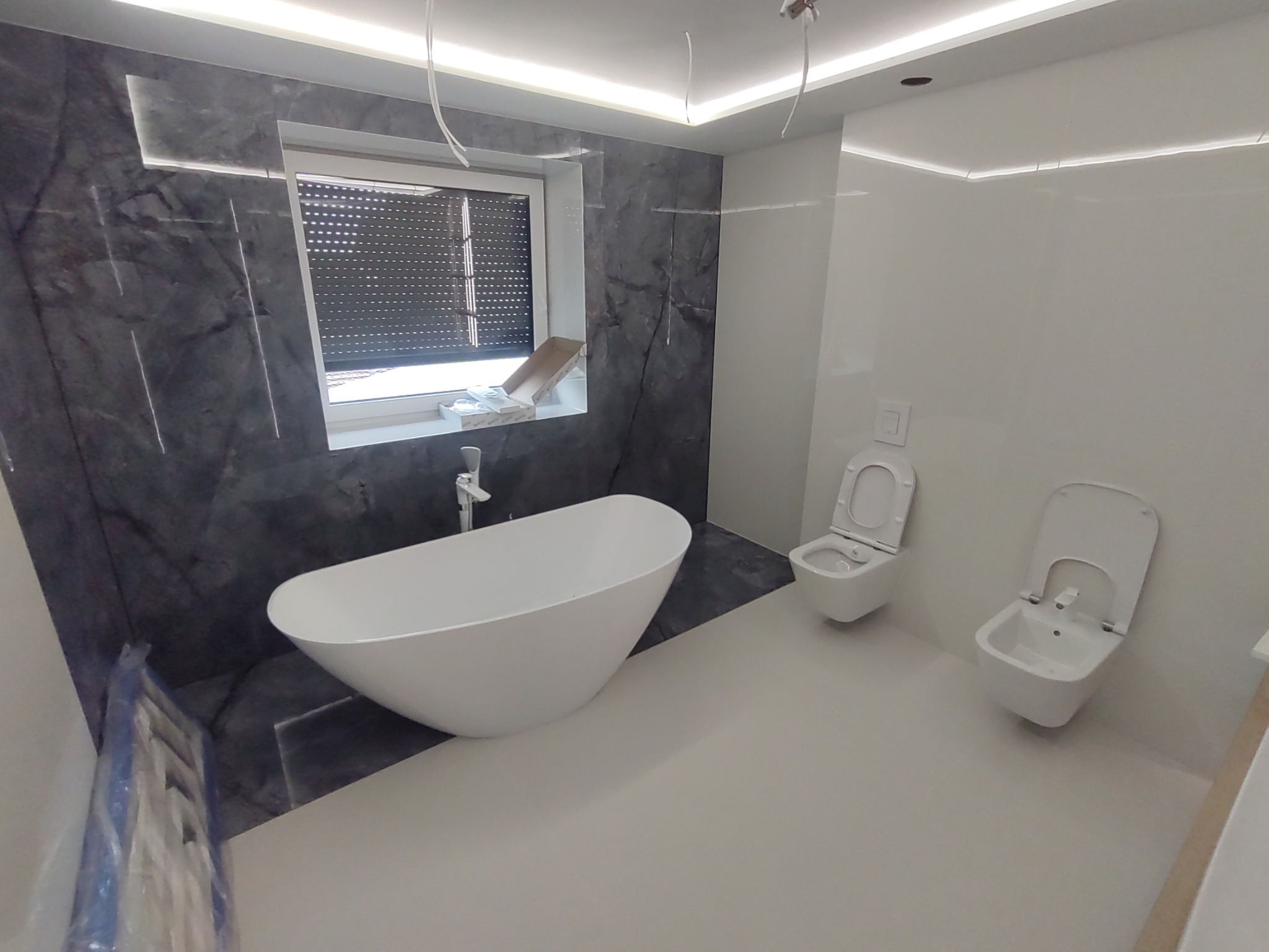 Nowoczesna łazienka z wolnostojącą wanną, marmurową ścianą, podwieszaną toaletą i bidetem, oświetleniem LED podsufitowym oraz oknem z żaluzjami.
