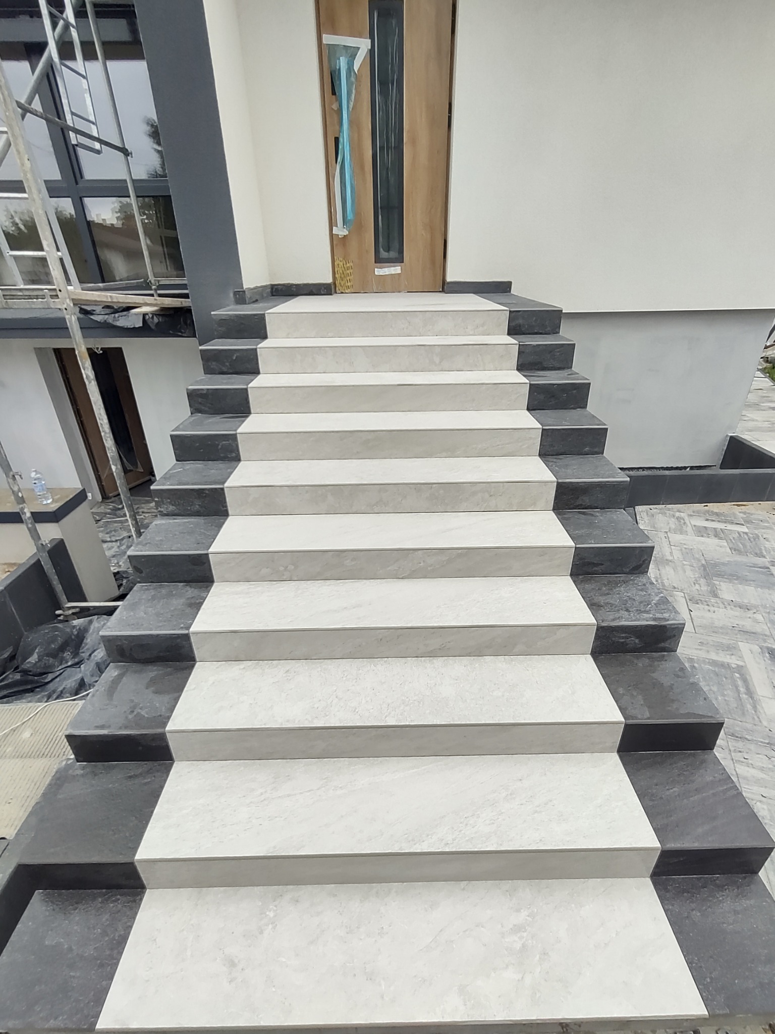 Zewnętrzne schody wejściowe wyłożone jasnoszarymi płytkami z ciemnoszarymi obrzeżami, prowadzące do drzwi wejściowych w nowym budynku, z widocznymi elementami rusztowania w tle.