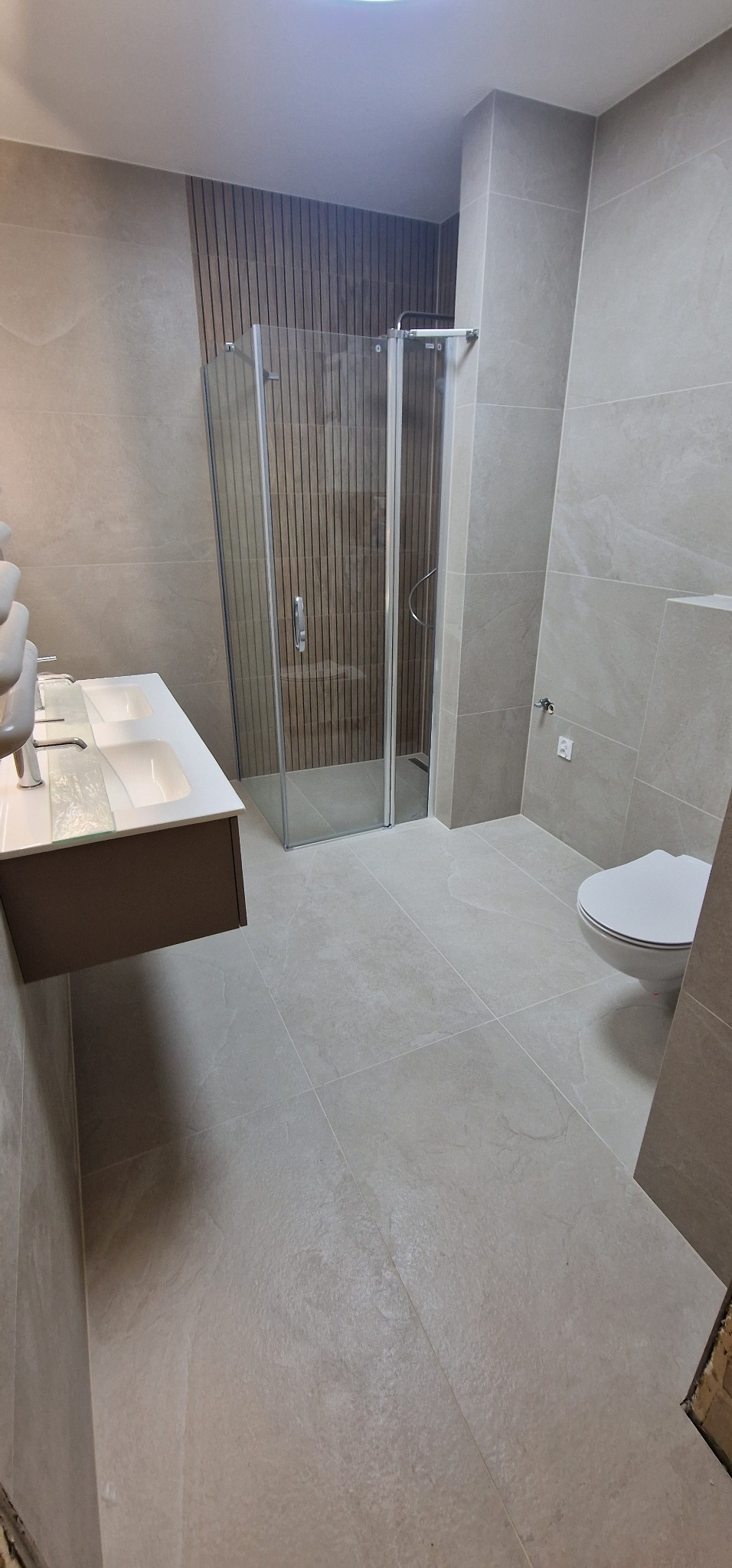 Nowoczesna łazienka z kabiną prysznicową, umywalką nablatową i toaletą, wykończona płytkami w odcieniach beżu i brązu z akcentem drewnianych paneli na ścianie prysznica.