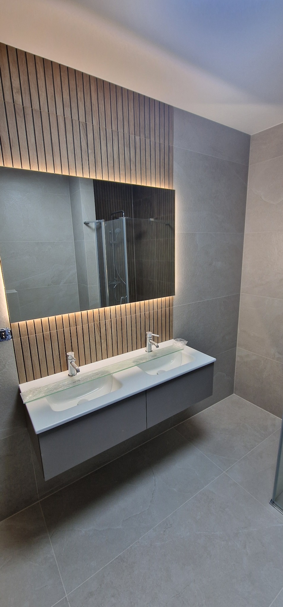 Nowoczesna łazienka z podwójną umywalką nablatową na szarej szafce, podświetlane lustro na tle drewnianej ściany z pionowych listew, szare płytki podłogowe i ścienne.