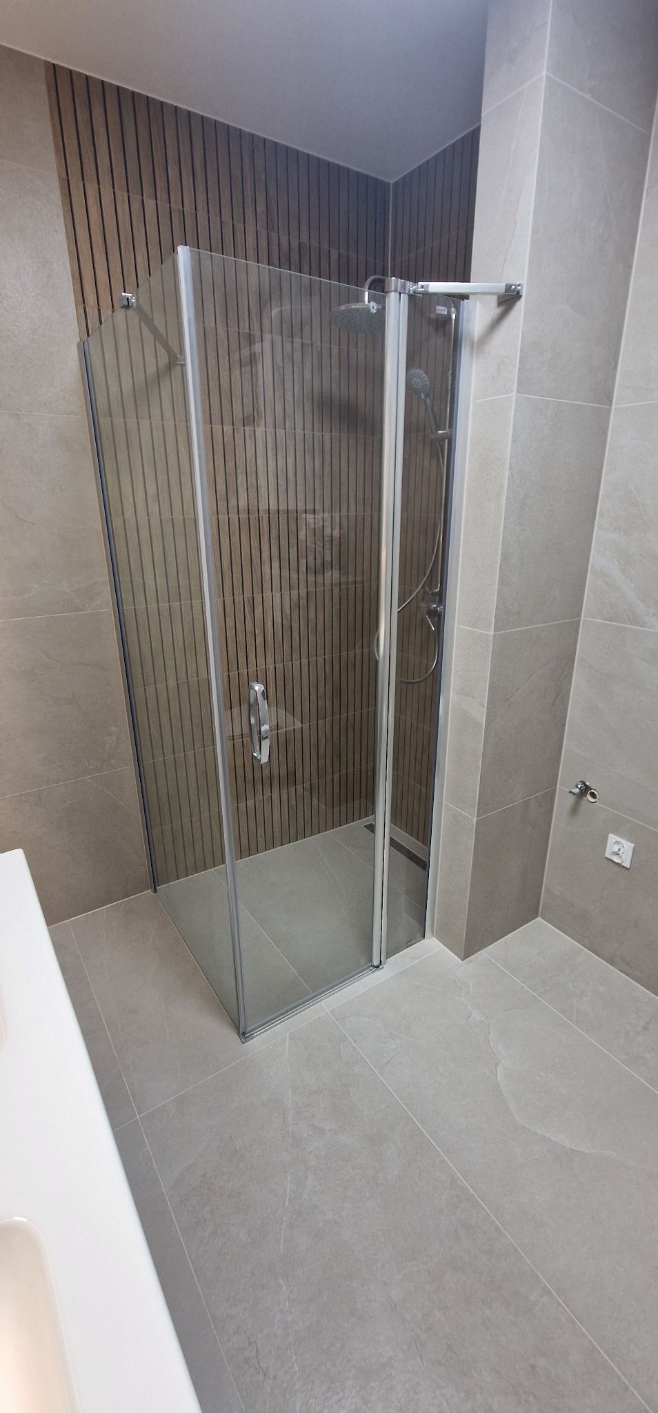 Nowoczesna kabina prysznicowa z szarymi płytkami imitującymi kamień, drewnopodobne lamele na ścianie i biała umywalka w rogu kadru, w łazience po remoncie.