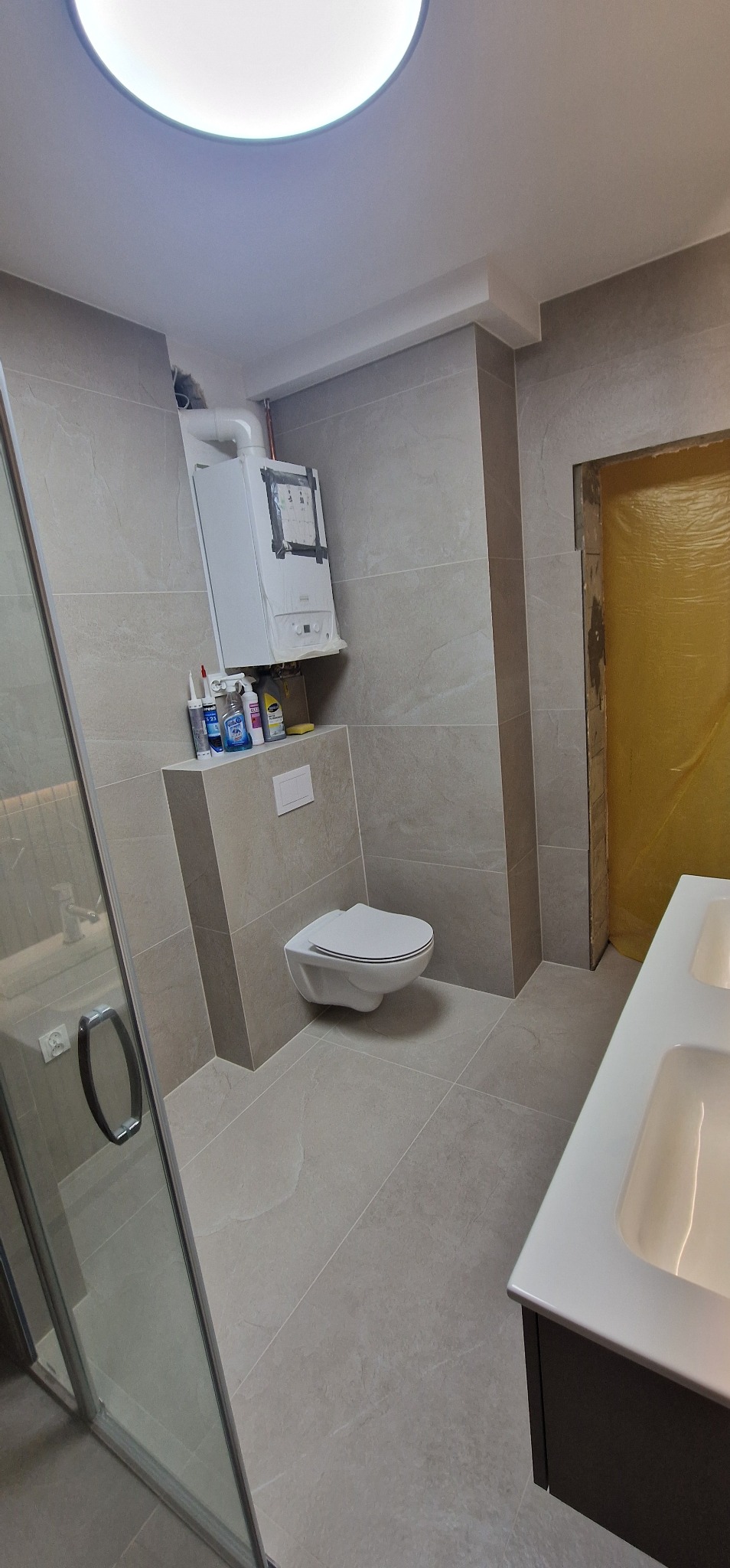 Wykończona łazienka z beżowymi płytkami, wiszącą toaletą i piecem gazowym, widok na kabinę prysznicową i umywalkę.