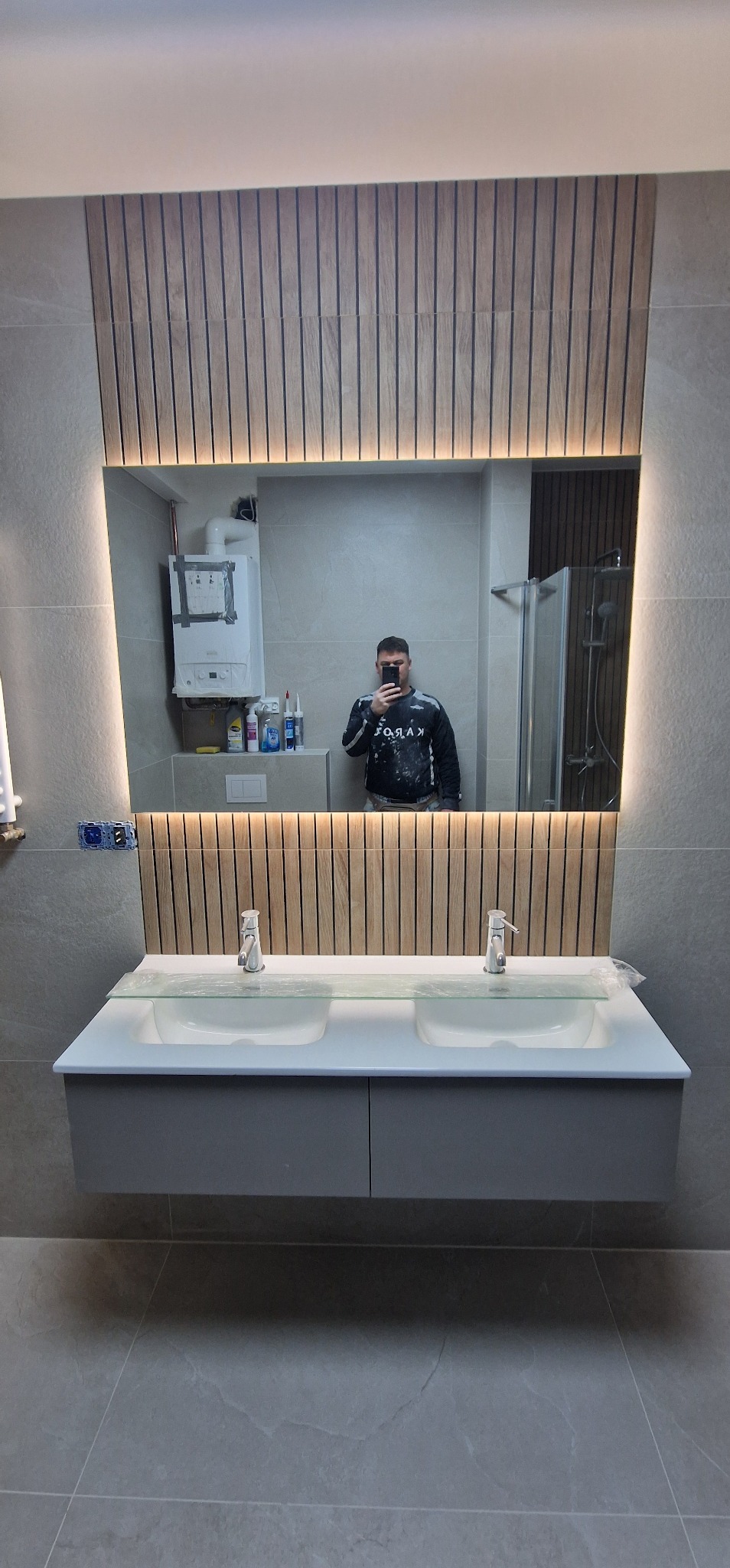 Nowoczesna łazienka z podwójną umywalką, lustrem z podświetleniem LED i drewnianymi panelami. W lustrze odbija się osoba robiąca zdjęcie oraz fragment kotła gazowego.