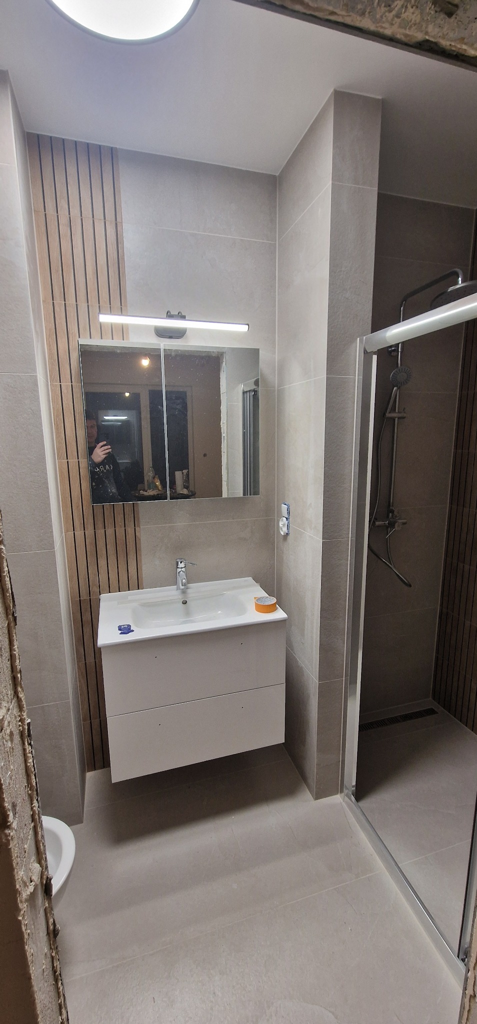 Nowoczesna łazienka w trakcie wykańczania: widoczna umywalka z szafką, lustro z oświetleniem, kabina prysznicowa z deszczownicą, płytki w odcieniach szarości, fragment ściany z drewnianymi panelami...