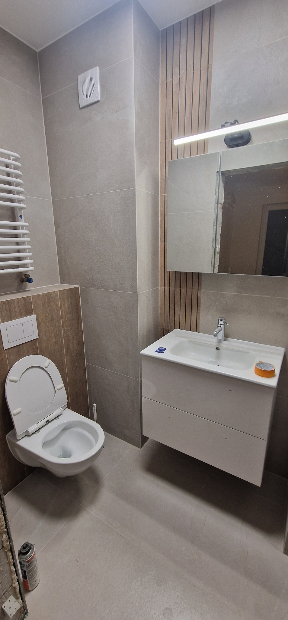 Nowoczesna łazienka w odcieniach szarości, z podwieszaną szafką umywalkową, lustrem z oświetleniem LED i toaletą z deską wolnoopadającą; widoczna puszka pianki montażowej przy podłodze.