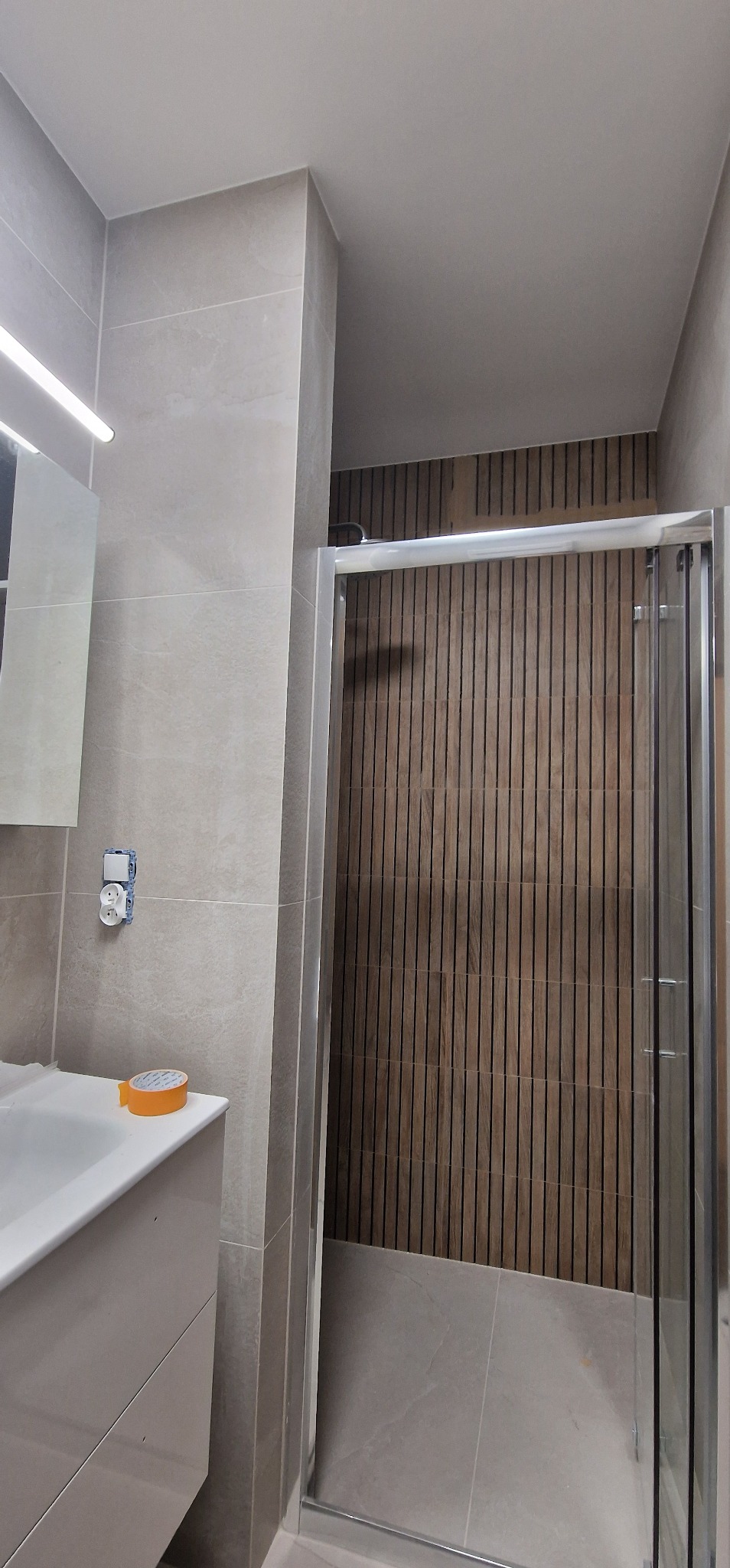 Wykończona łazienka z kabiną prysznicową z drewnianymi panelami na ścianie, szare płytki na ścianach i podłodze oraz biała umywalka z szafką. Widoczna taśma malarska na umywalce i gniazdko...