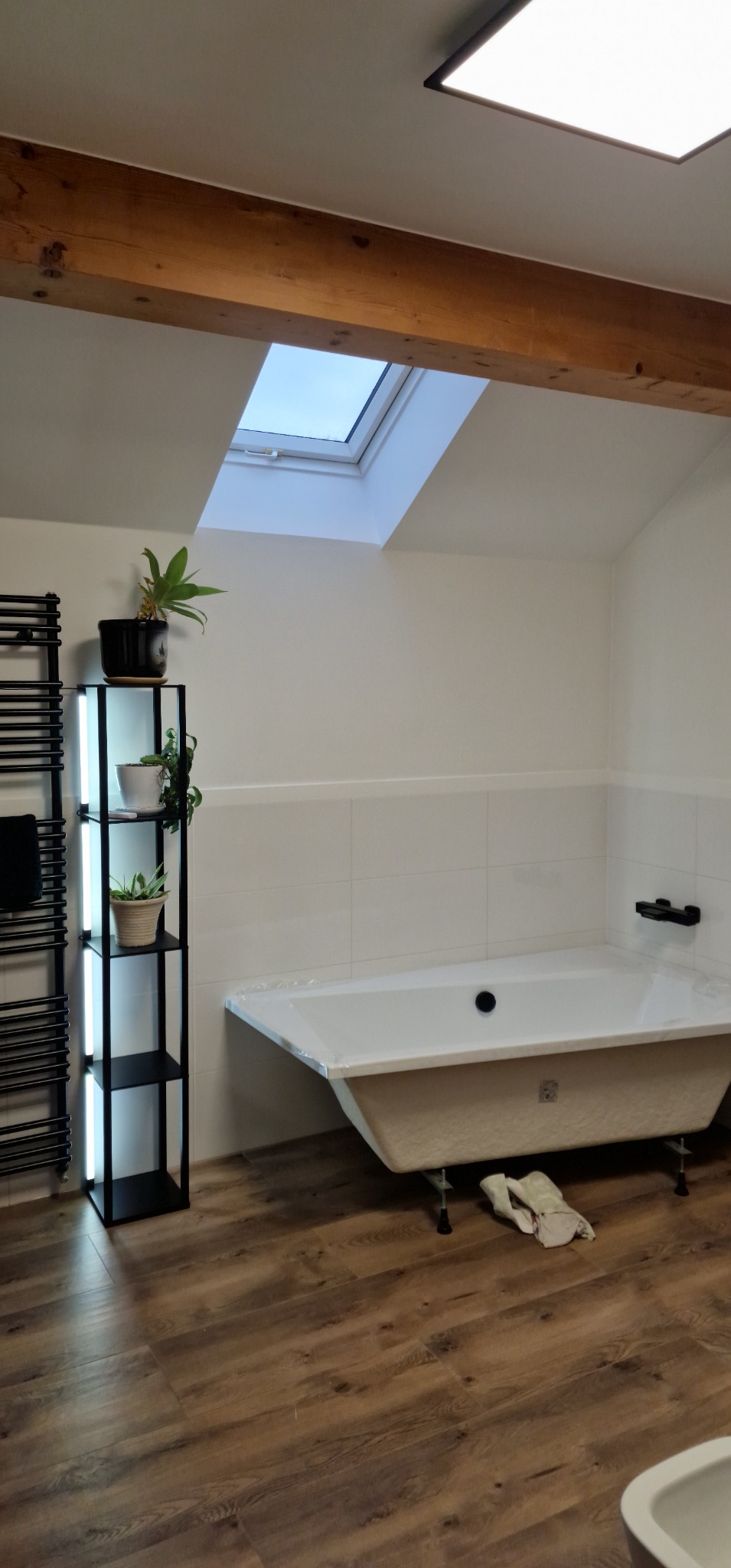 Jasna łazienka na poddaszu z drewnianą belką stropową, wanną na nóżkach, grzejnikiem drabinkowym i półką z roślinami doniczkowymi, oświetlona światłem z okna dachowego.