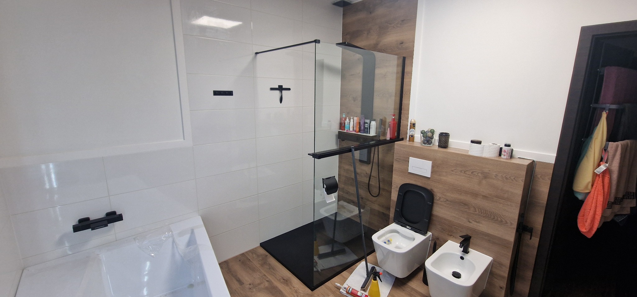 Nowoczesna łazienka z kabiną prysznicową typu walk-in z czarnymi detalami, białą armaturą, obudową z paneli imitujących drewno i częściowo wykończona białymi płytkami. Widoczne narzędzia...