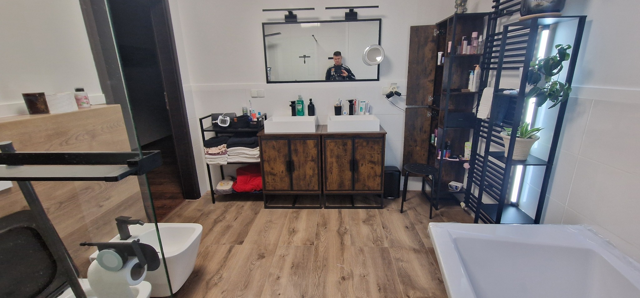 Nowoczesna łazienka z drewnopodobną podłogą, podwójną umywalką z szafką w stylu industrialnym, regałem na kosmetyki i rośliny oraz lustrem z odbijającą się osobą.