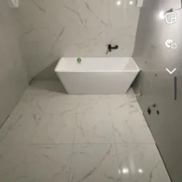 Nowoczesna łazienka z białą, trapezoidalną wanną, marmurowymi płytkami na ścianach i podłodze, oraz czarną armaturą. Widoczne niedokończone prace instalacyjne na ścianie.