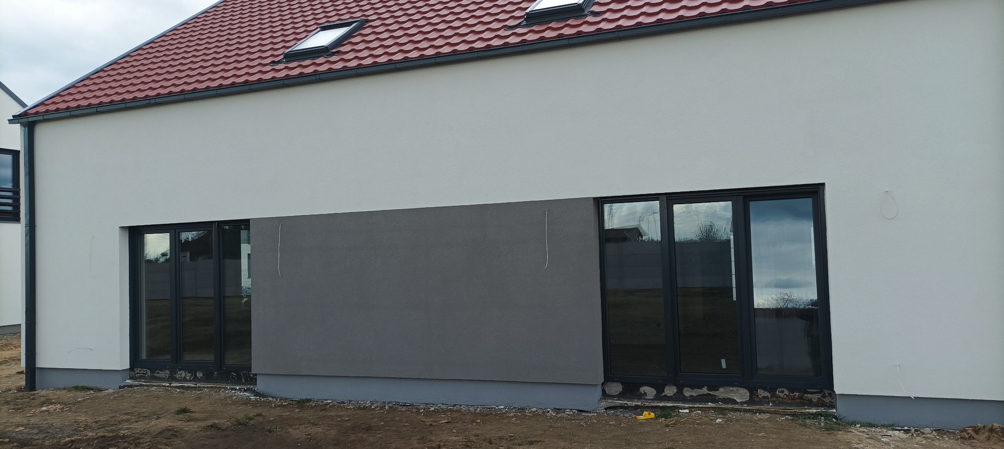 Świeżo otynkowana elewacja domu jednorodzinnego z widocznymi oknami z czarnymi ramami. Dolna część elewacji w kolorze szarym, górna biała. Widoczna czerwona dachówka na dachu.