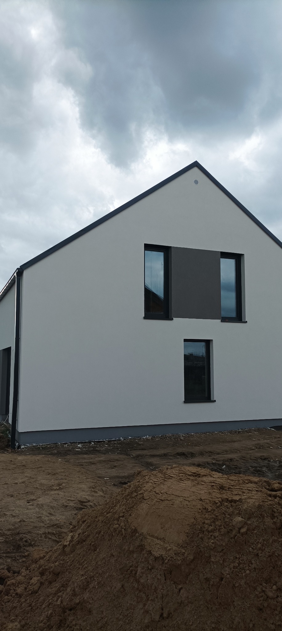 Nowoczesny dom z jasną elewacją i szarym akcentem między oknami, widoczne ślady prac ziemnych wokół budynku.