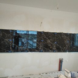 Usługi remontowo budowlane Mykola Dmytryk - Montaż płytek imitujących marmur na ścianie, widoczne niebieskie kliny dystansowe i odbicie osoby w jednej z płytek.