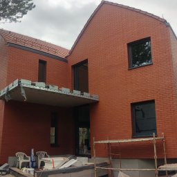 Usługi remontowo budowlane Mykola Dmytryk - Nowoczesny dom z czerwonej cegły z ciemnymi oknami, w trakcie budowy, z widocznym rusztowaniem i materiałami budowlanymi na pierwszym planie.