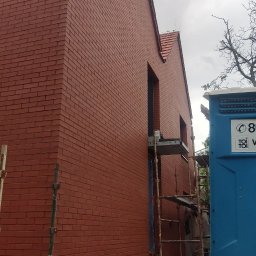 Usługi remontowo budowlane Mykola Dmytryk - Nowo wybudowany budynek z cegły klinkierowej w odcieniach czerwieni, widoczne rusztowania i przenośna toaleta na placu budowy, pochmurne niebo w tle.