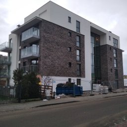 Usługi remontowo budowlane Mykola Dmytryk - Nowoczesny budynek wielorodzinny z ceglaną elewacją i balkonami, widoczny kontener na gruz i materiały budowlane, sugerujący trwające prace wykończeniowe.