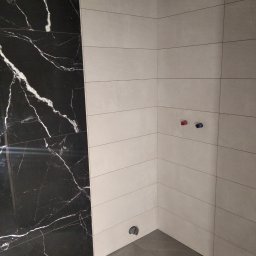 Usługi remontowo budowlane Mykola Dmytryk - Fragment łazienki z wyłożonymi płytkami: ściana z czarnych płytek imitujących marmur i ściana z beżowych, poziomych płytek. Widoczne przyłącza wodne i odpływ pod prysznic.