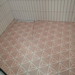 Usługi remontowo budowlane Mykola Dmytryk - Wykończona łazienka z jasnoróżowymi, heksagonalnymi płytkami podłogowymi z geometrycznym wzorem i wąskimi, pionowymi białymi płytkami ściennymi z czarnymi fugami.