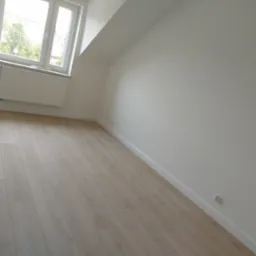 Jasny pokój na poddaszu z ułożonymi panelami podłogowymi, białe ściany i listwy przypodłogowe, okno z grzejnikiem.