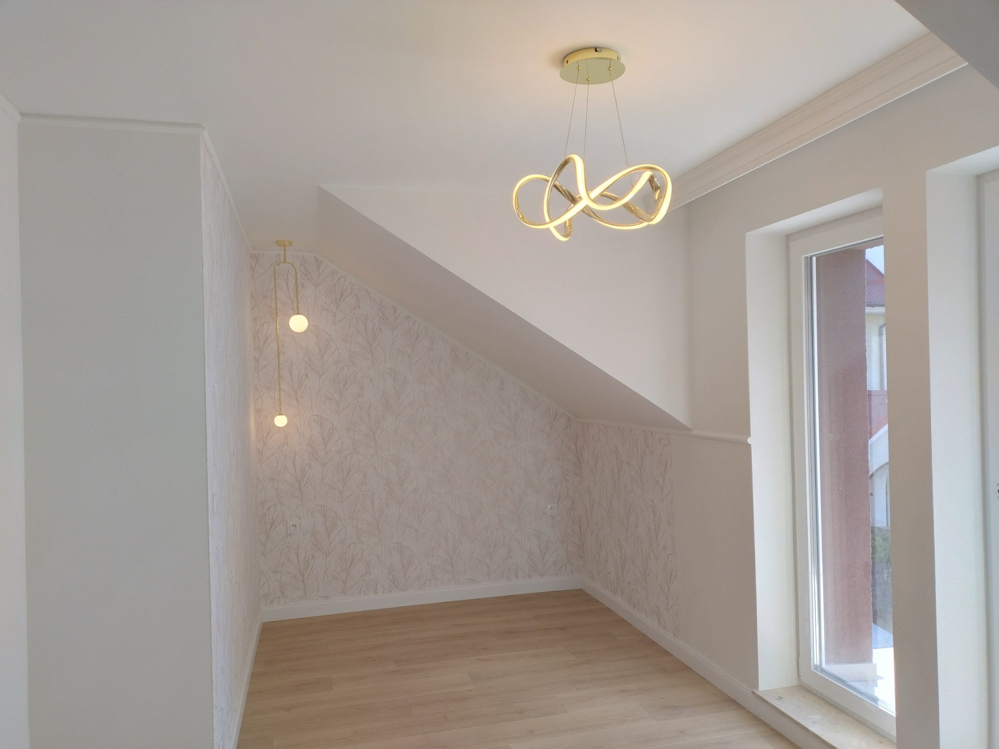 Poddasze po remoncie z jasnymi panelami, ścianami w pastelowych barwach i nowoczesnym oświetleniem: designerska lampa wisząca i minimalistyczna lampa ścienna.