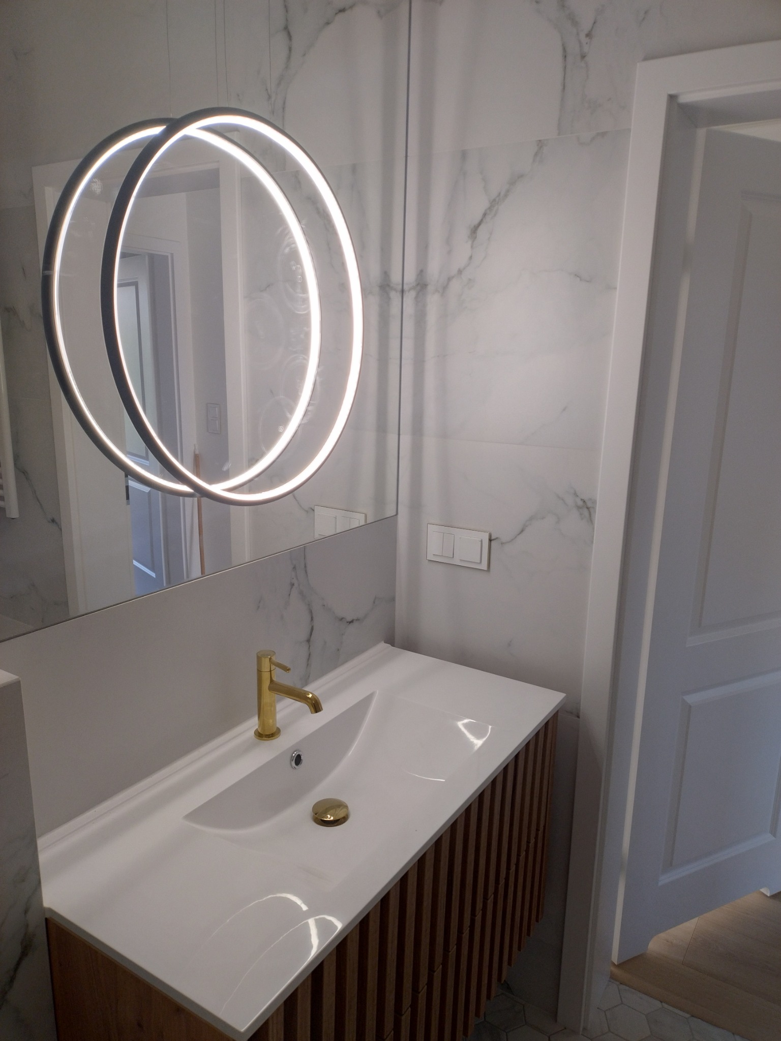 Nowoczesna łazienka z umywalką nablatową, złotą armaturą i okrągłym lustrem z podświetleniem LED na tle marmurowej ściany.