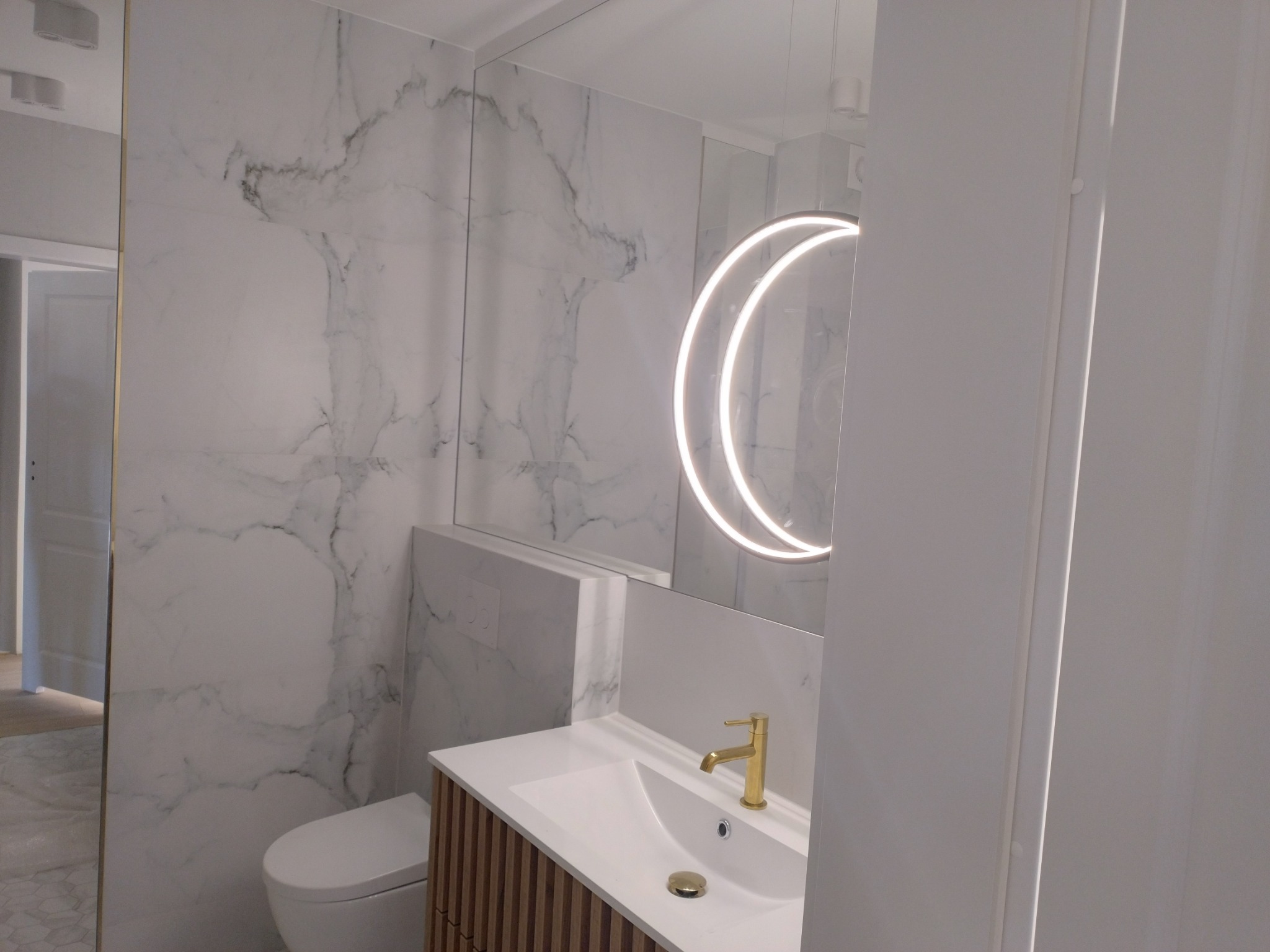 Nowoczesna łazienka z marmurowymi płytkami, podświetlanym lustrem w kształcie półksiężyca i złotą armaturą, widoczna toaleta i umywalka z szafką o drewnianej fakturze.