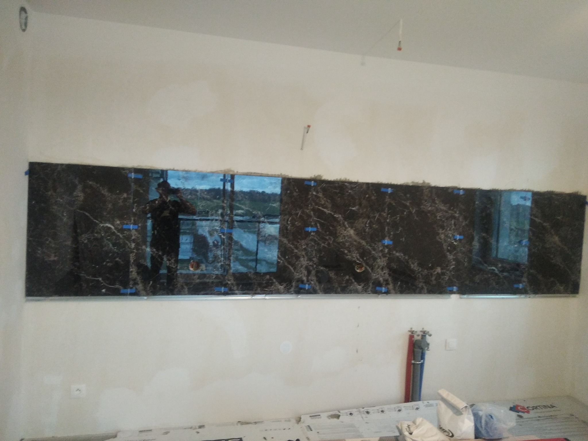 Montaż płytek imitujących marmur na ścianie, widoczne niebieskie kliny dystansowe i odbicie osoby w jednej z płytek.