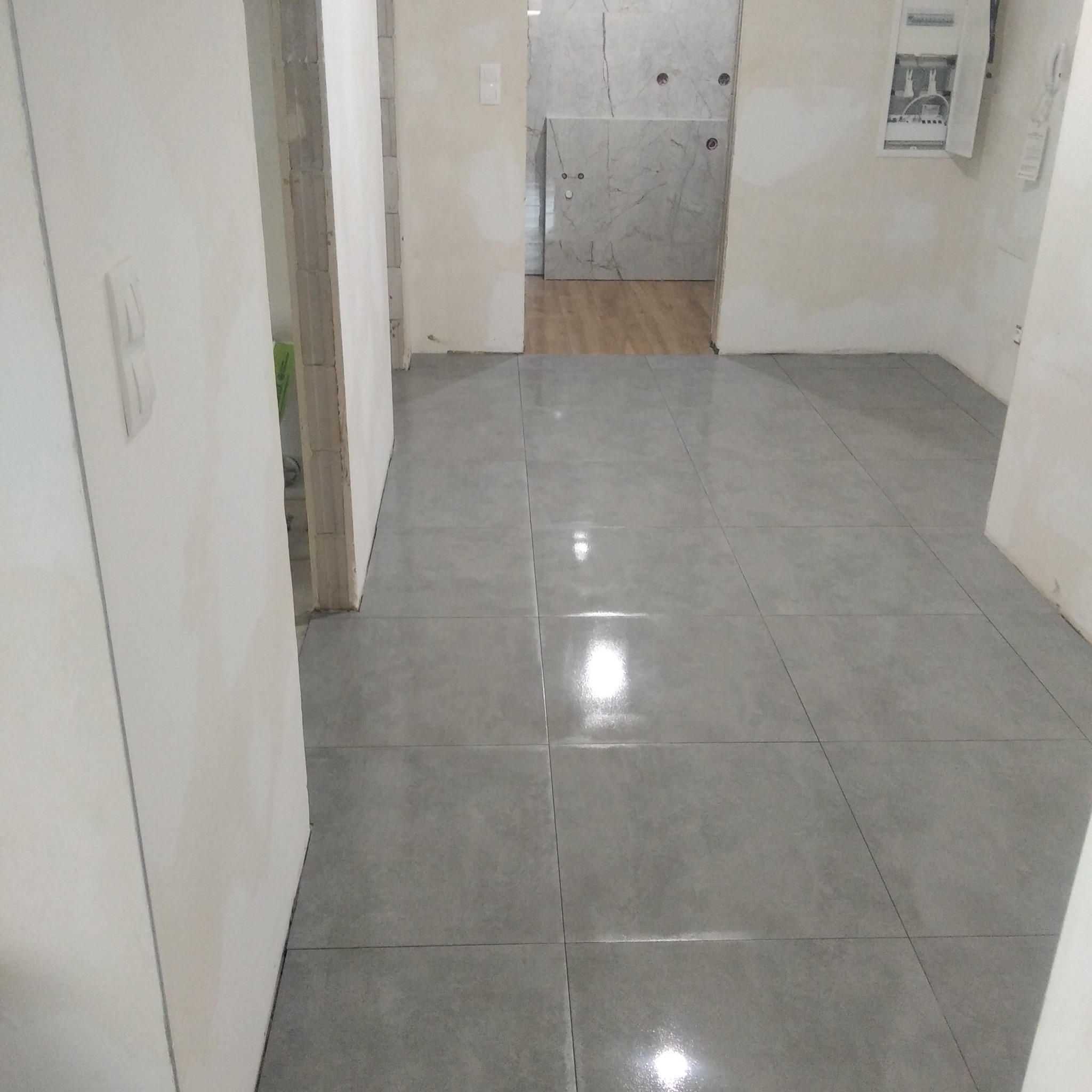 Ułożone szare płytki podłogowe w korytarzu, widok na łazienkę z marmurowymi płytkami i drewnianą podłogą, ściany przygotowane do malowania, widoczna skrzynka elektryczna.