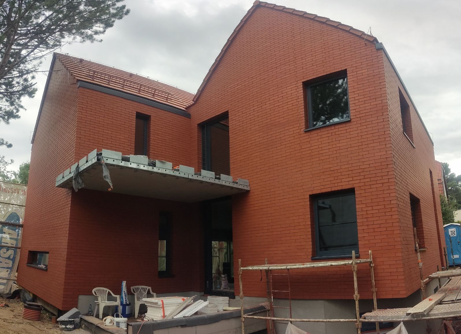 Nowoczesny dom z czerwonej cegły z ciemnymi oknami, w trakcie budowy, z widocznym rusztowaniem i materiałami budowlanymi na pierwszym planie.