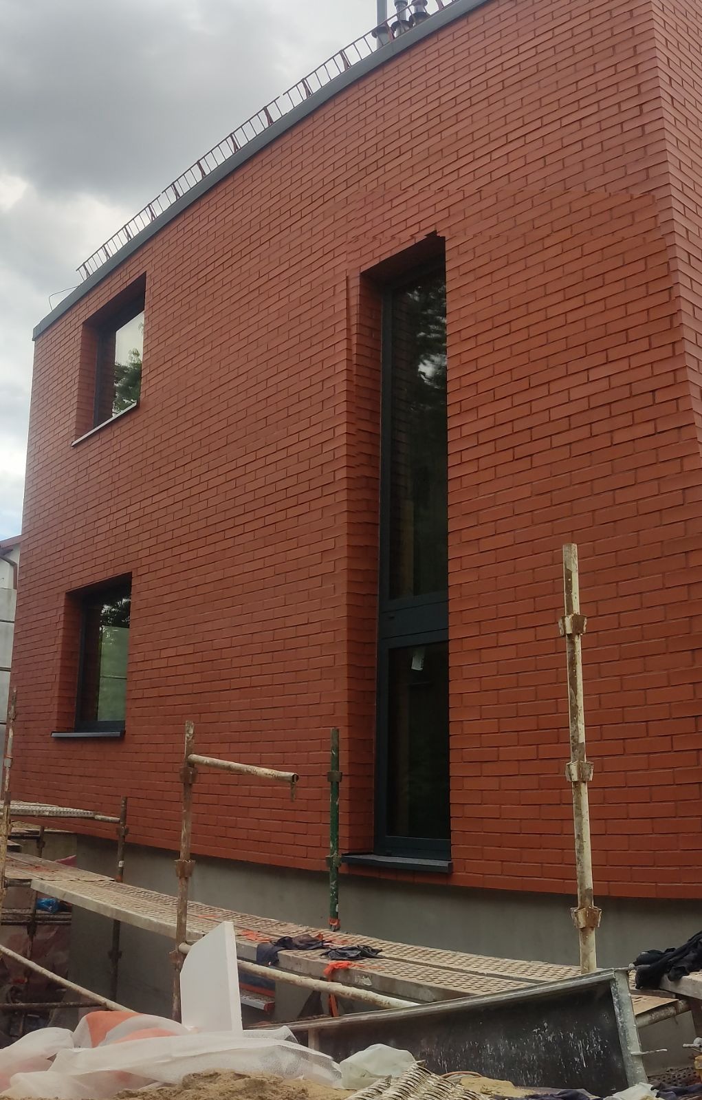 Czerwona ceglana elewacja budynku z zamontowanymi oknami, widoczne rusztowania i materiały budowlane w trakcie prac wykończeniowych.